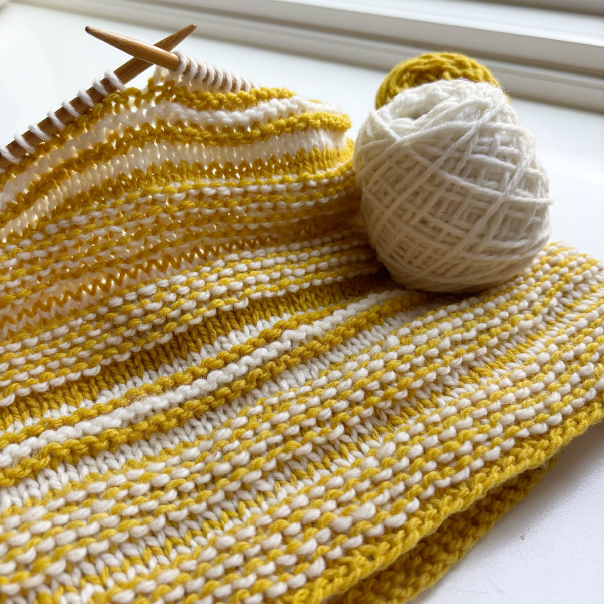 A two-color garter stitch cowl in gold and off-white yarn, pictured on a circular needle.