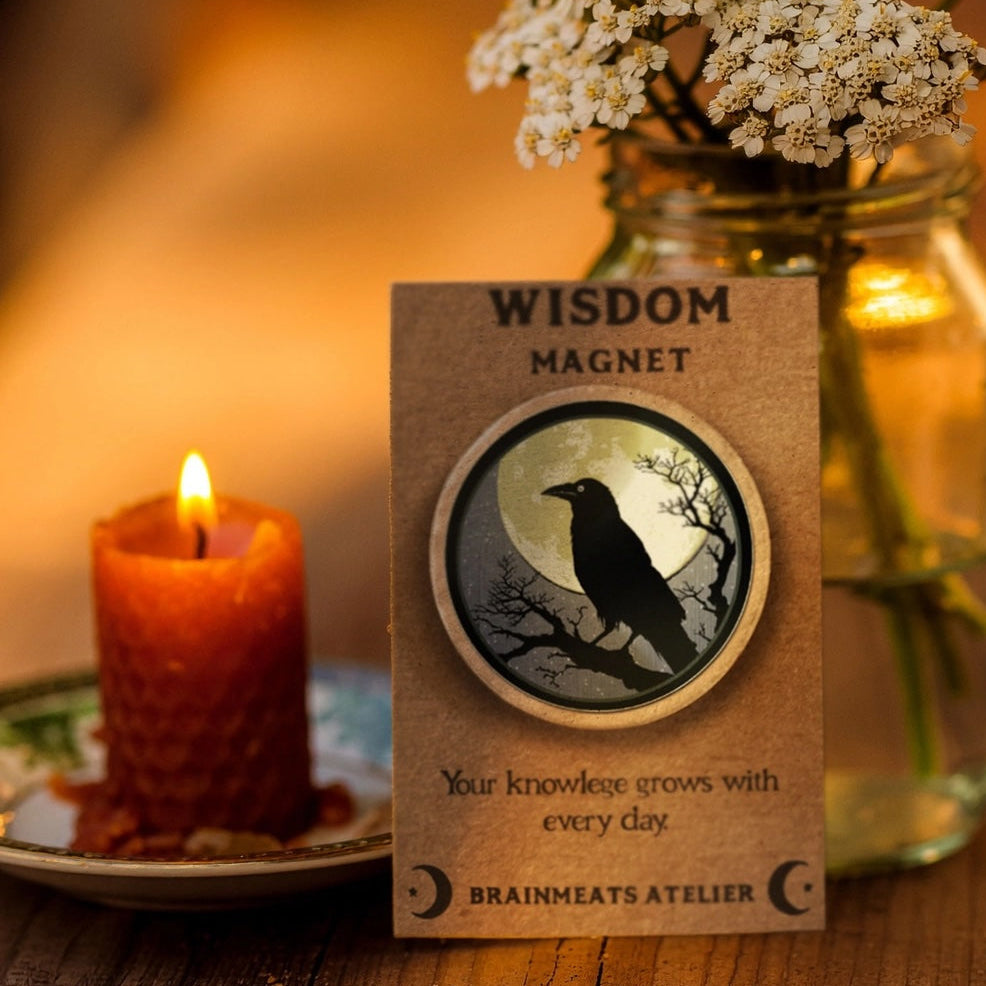 Wisdom magnet with a crow design next to a candle and flowers on a wooden surface