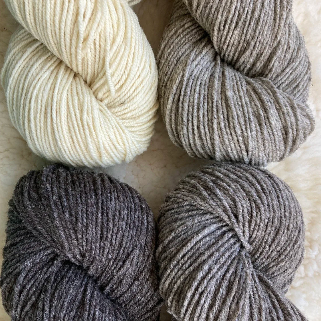 Four skeins of yarn in beige, gray, and black on a textured surface.