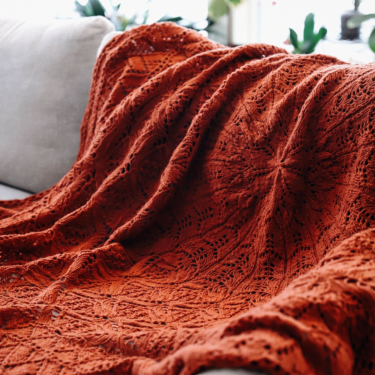A rust-orange open-knit crocheted throw blanket draped over a light beige sofa in a bright, softly lit room.