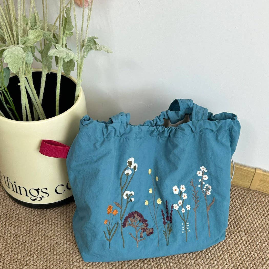 A blue fabric drawstring tote bag with embroidered wildflowers, placed on a woven beige rug next to a cream-colored ceramic planter with artificial flowers.