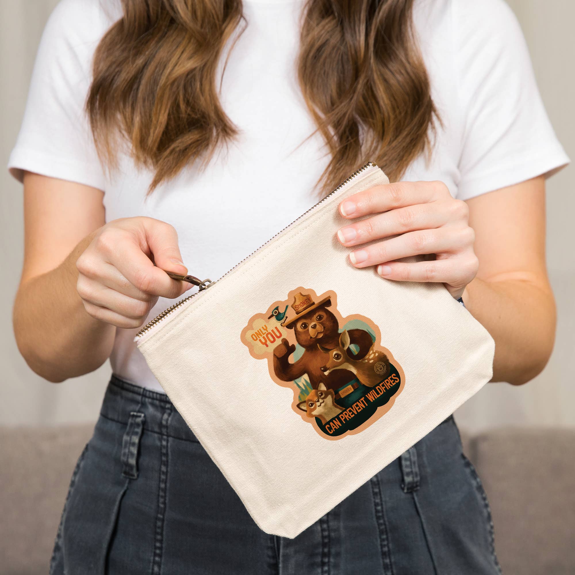 A person in a white t-shirt and dark jeans holds a cream canvas zipper pouch featuring a cartoon graphic of a bearded man with a camera and the text 'YOU CAN'T PROUGH WOLFIES'.