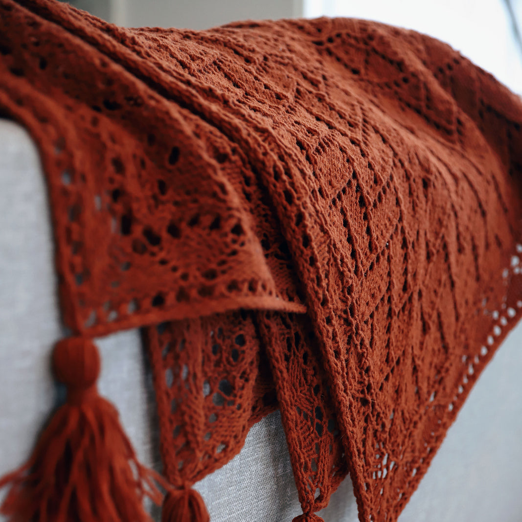 A rust-orange crocheted throw blanket with tassel ends draped over a light gray sofa in a softly lit interior setting.