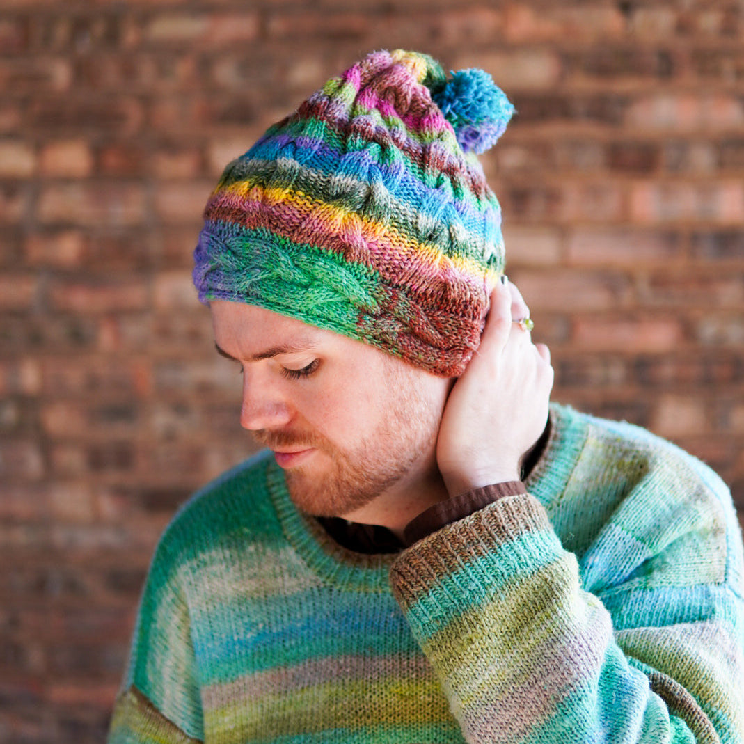A man wearing a colorful striped knit beanie with a pom-pom and a matching striped knit sweater, standing against a brick wall.