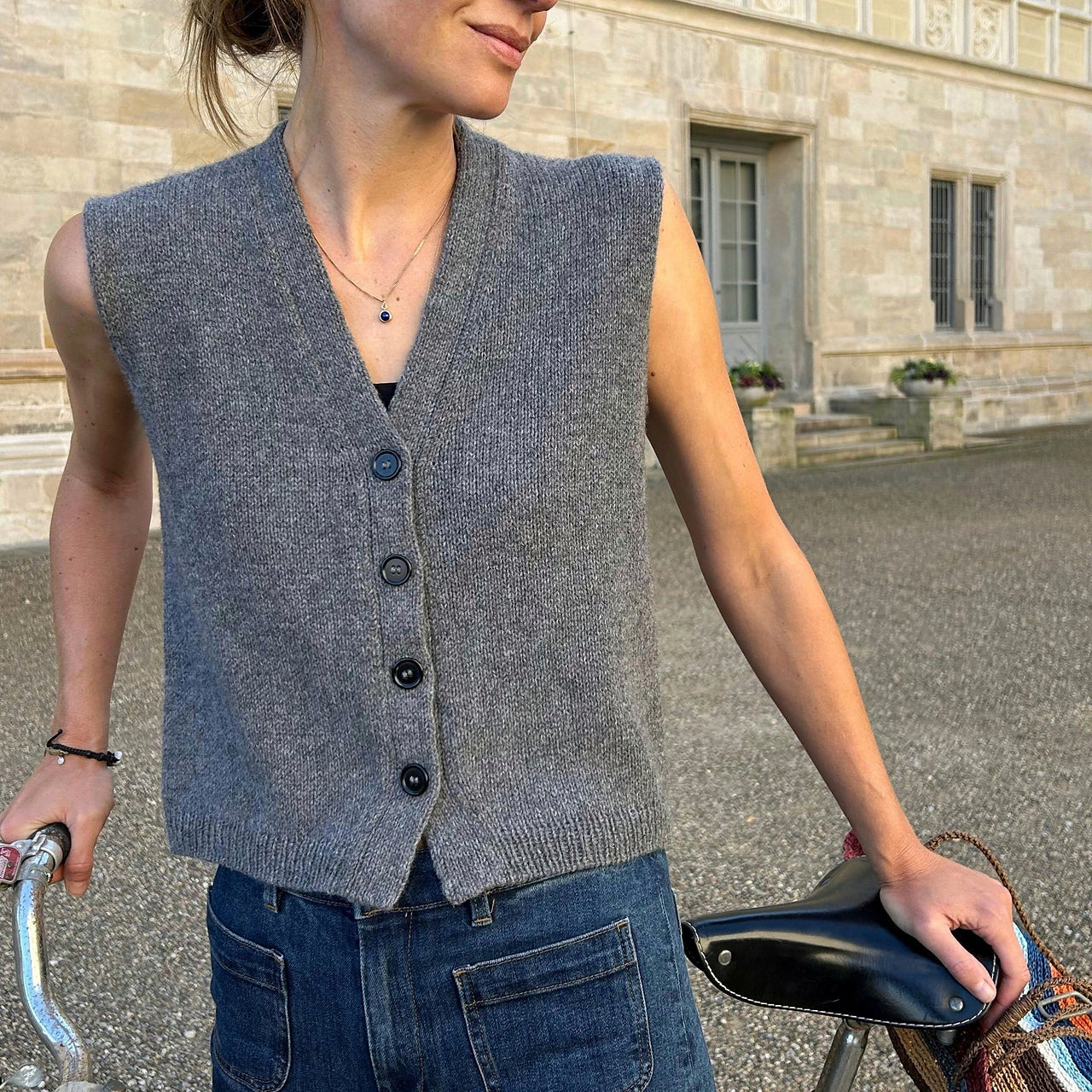 Person wearing a gray sleeveless sweater vest and blue jeans, holding a bicycle outdoors.