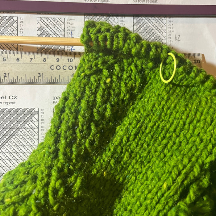 a partly knit green sweater with a patterned band rests on top of a knitting chart.