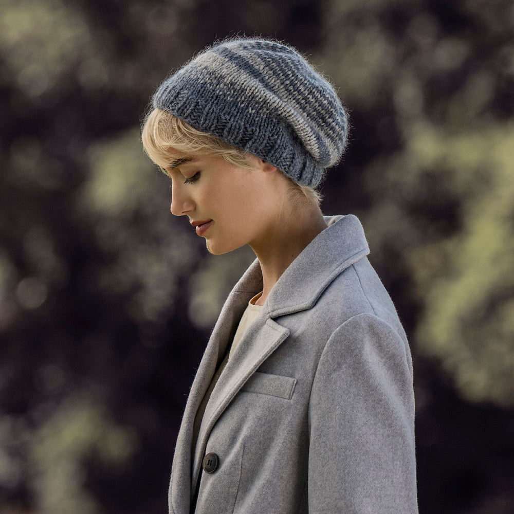 A woman in profile wearing a chunky knit beanie hat in heathered blue-gray and navy stripes, paired with a light gray coat, set against a soft, blurred outdoor background.