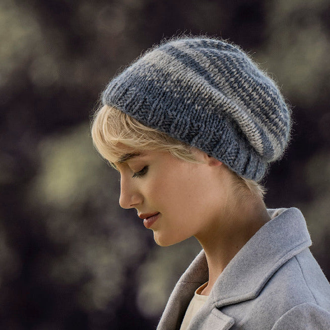 A woman in profile wearing a chunky knit beanie hat in heathered blue-gray and navy stripes, paired with a light gray coat, set against a soft, blurred outdoor background.