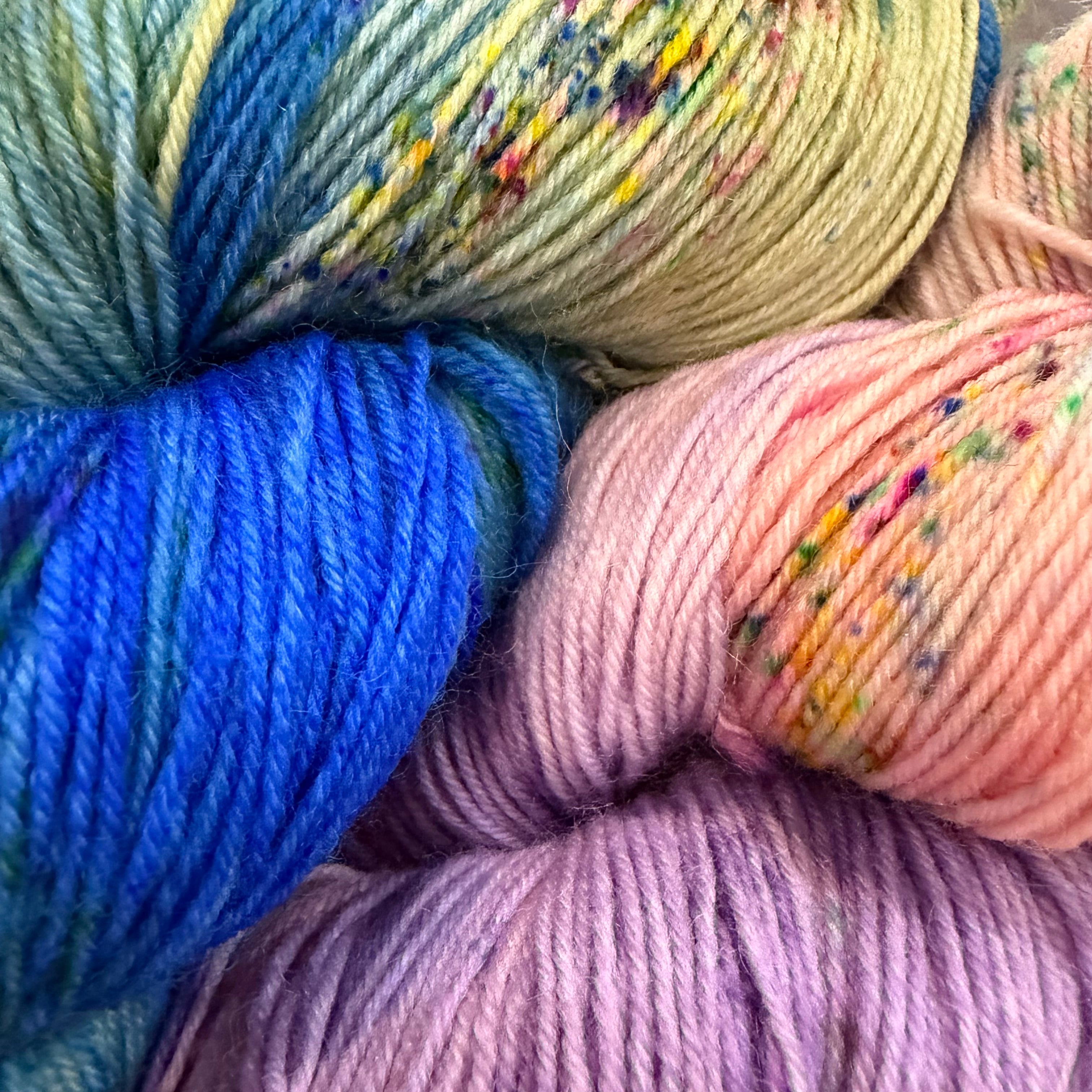 Close-up of several skeins of variegated yarn in pastel and bright tones, including blue, yellow, pink, and lavender, with small multicolor flecks throughout.