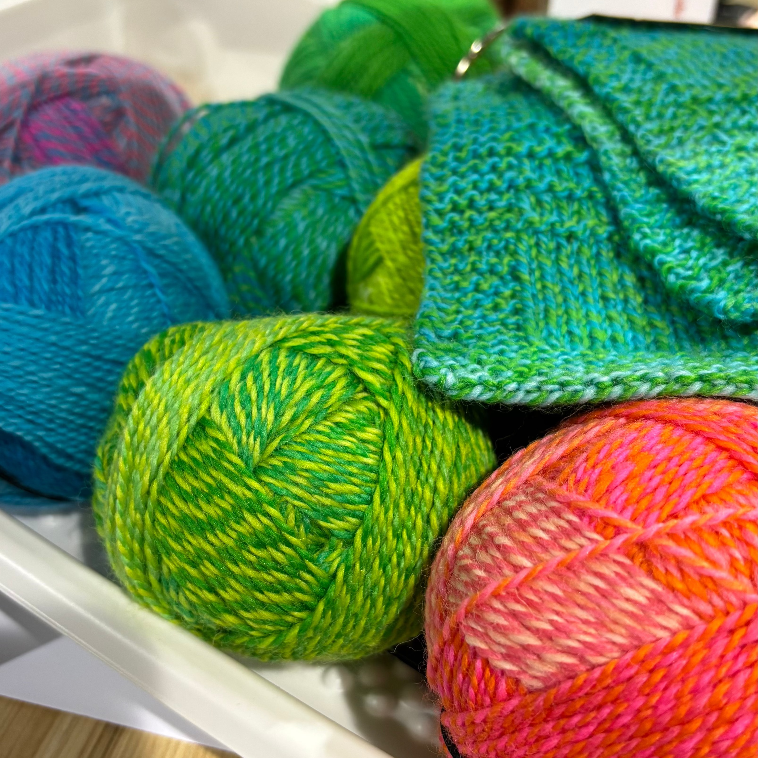 A close-up of colorful variegated yarn balls and a matching knitted fabric sample displayed in a white tray on a wooden table.