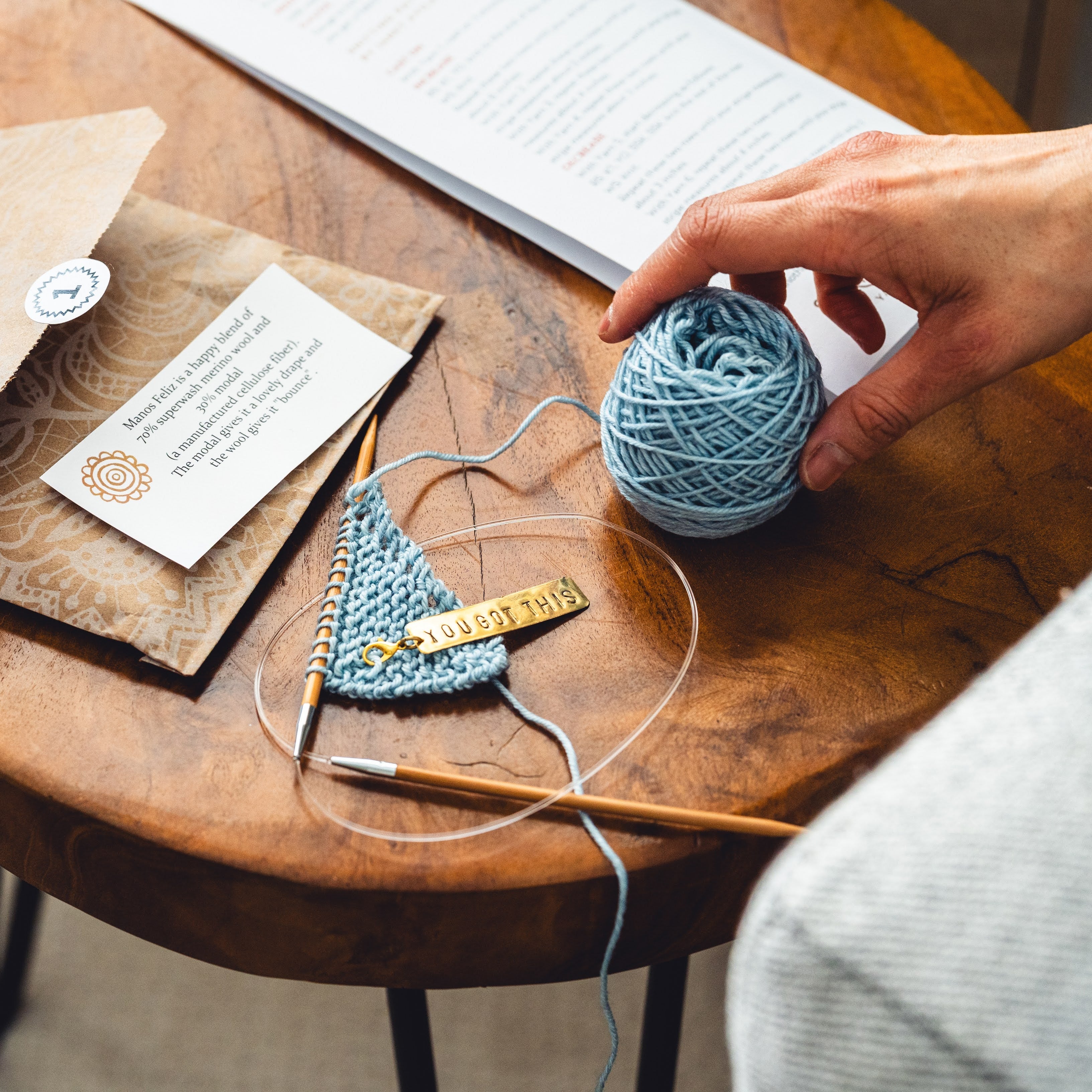  a hand reaching for a ball of yarn that is sitting on a table with a Box of Light envelope and the start of a project on the needles.