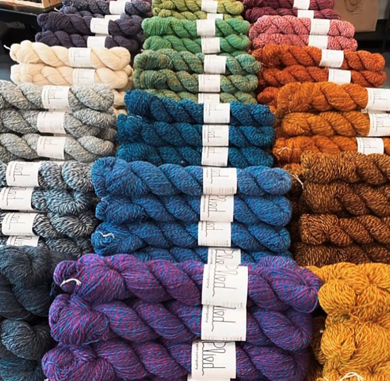 A close-up display of multiple skeins of yarn arranged in rows, showing a wide range of colors including blue, purple, green, orange, brown, and cream. Each skein has a small white paper label wrapped around it.