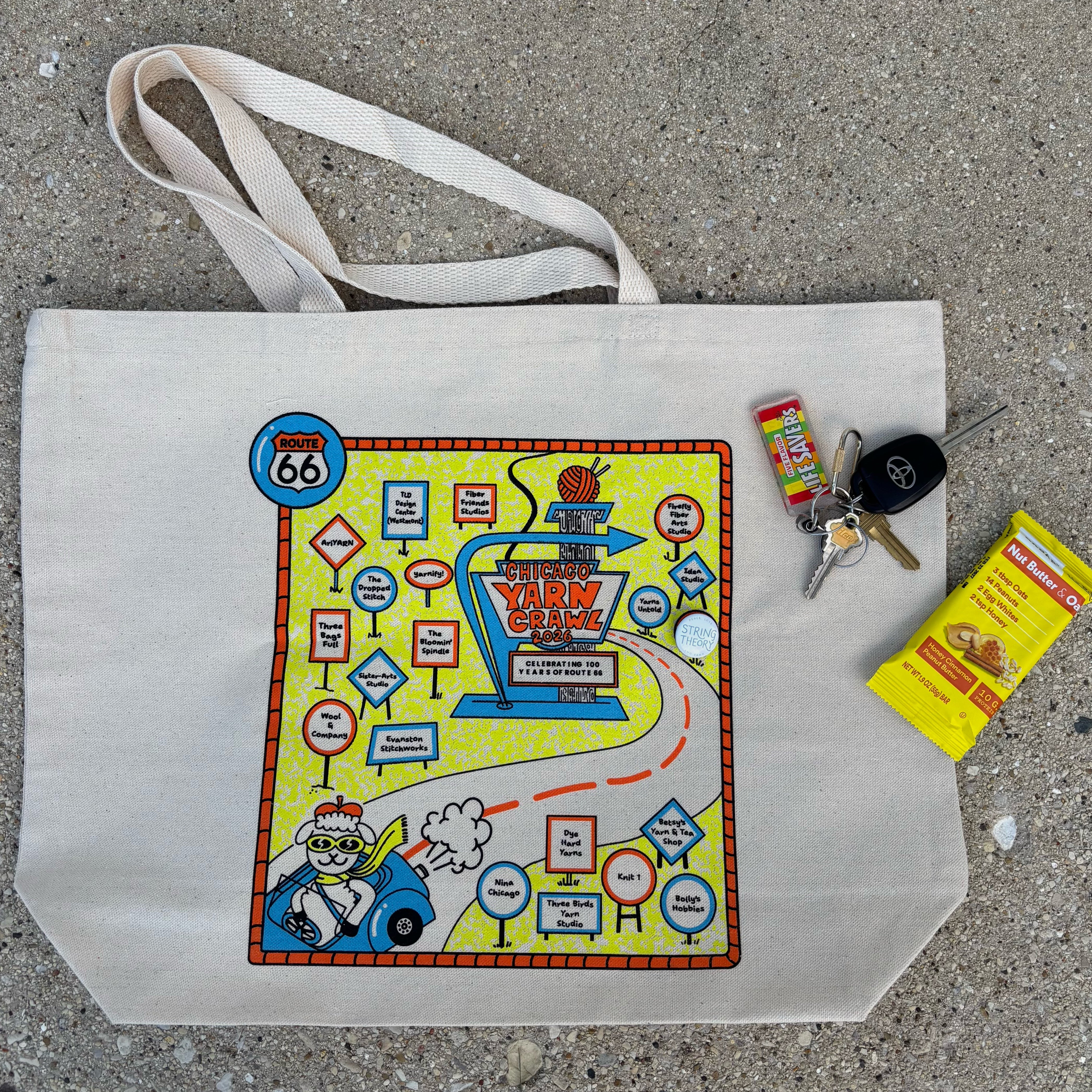 A beige canvas tote bag laid flat on a gray concrete surface, featuring a colorful vintage-style map graphic with the words 'The Great Yarn Crawl' and Route 66. Small props including keys, and a protein bar.