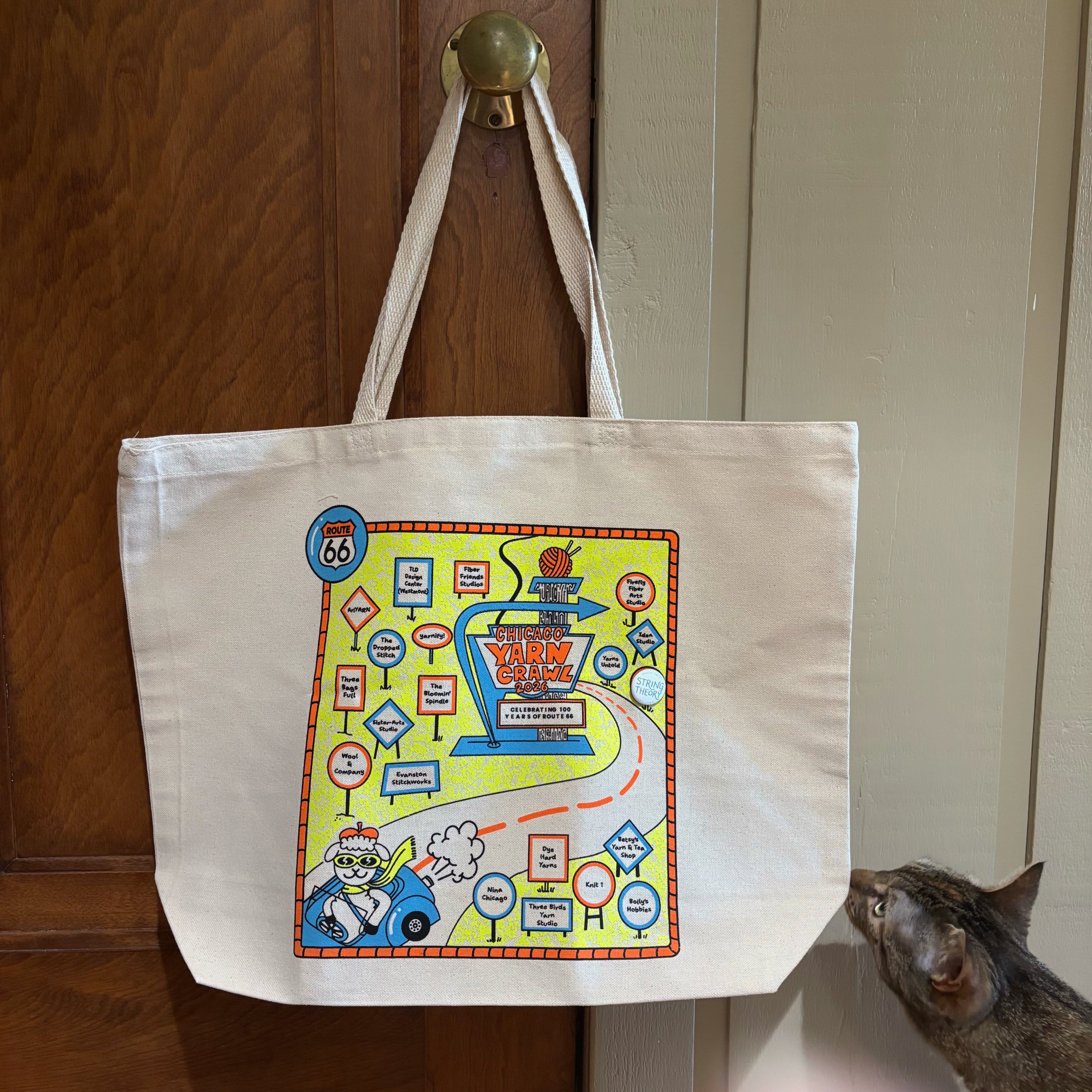 An off-white canvas tote bag with a colorful vintage-style map graphic hanging from a brass doorknob on a wooden door. A tabby cat is peeking in from the right side of the frame.