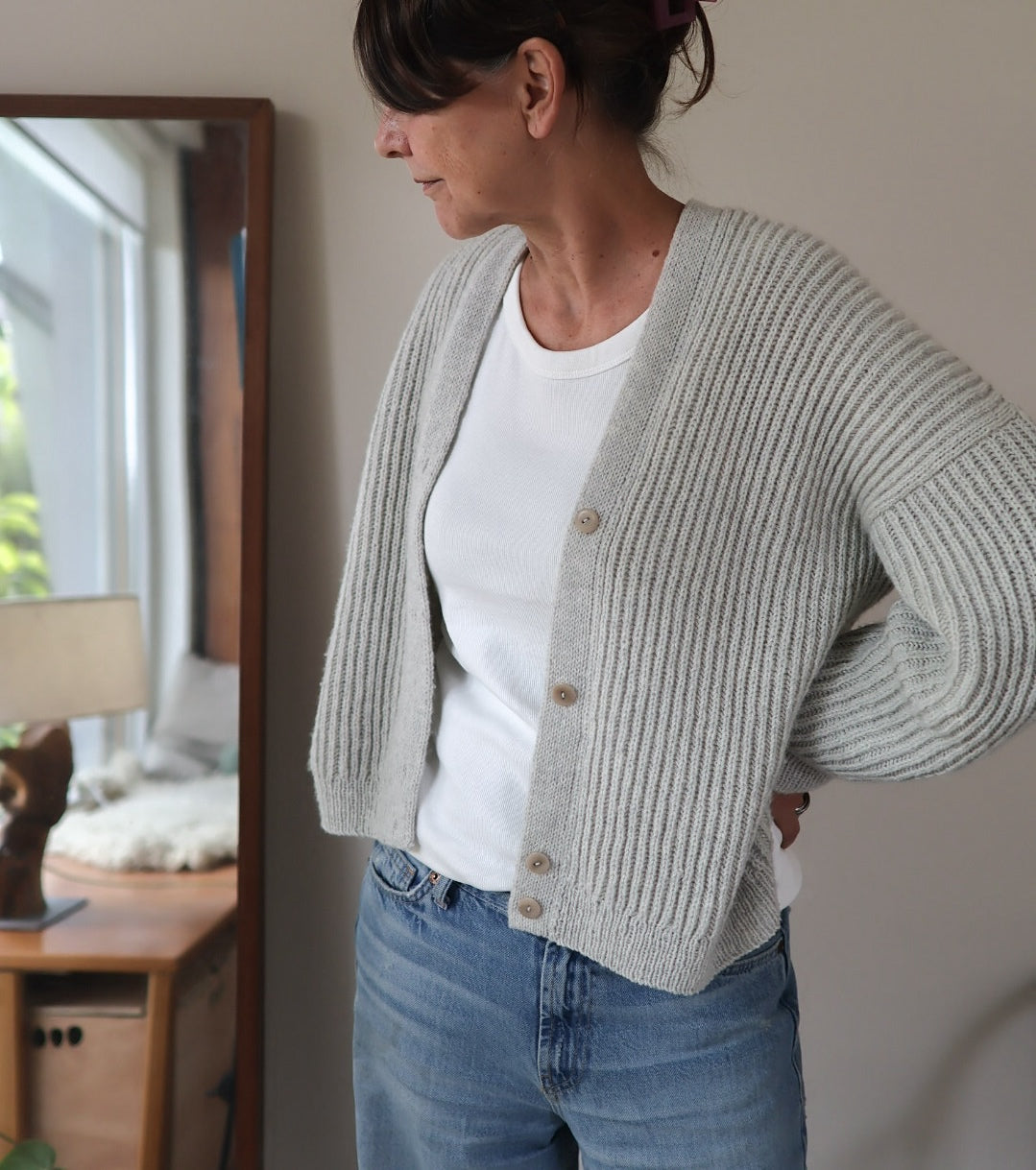 Person wearing a light gray knitted cardigan over a white shirt and blue jeans, standing indoors.